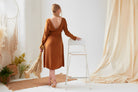 A woman in a Sarvin Mustard Yellow Midi Dress stands with her back to the camera, holding pampas grass and resting her hand on a white chair in a softly decorated, neutral-toned room.