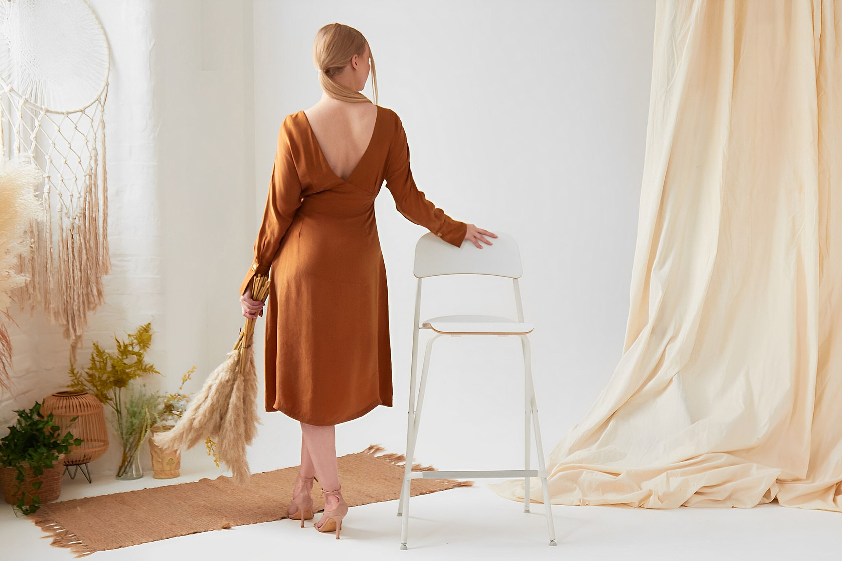 A woman in a Sarvin Mustard Yellow Midi Dress stands with her back to the camera, holding pampas grass and resting her hand on a white chair in a softly decorated, neutral-toned room.