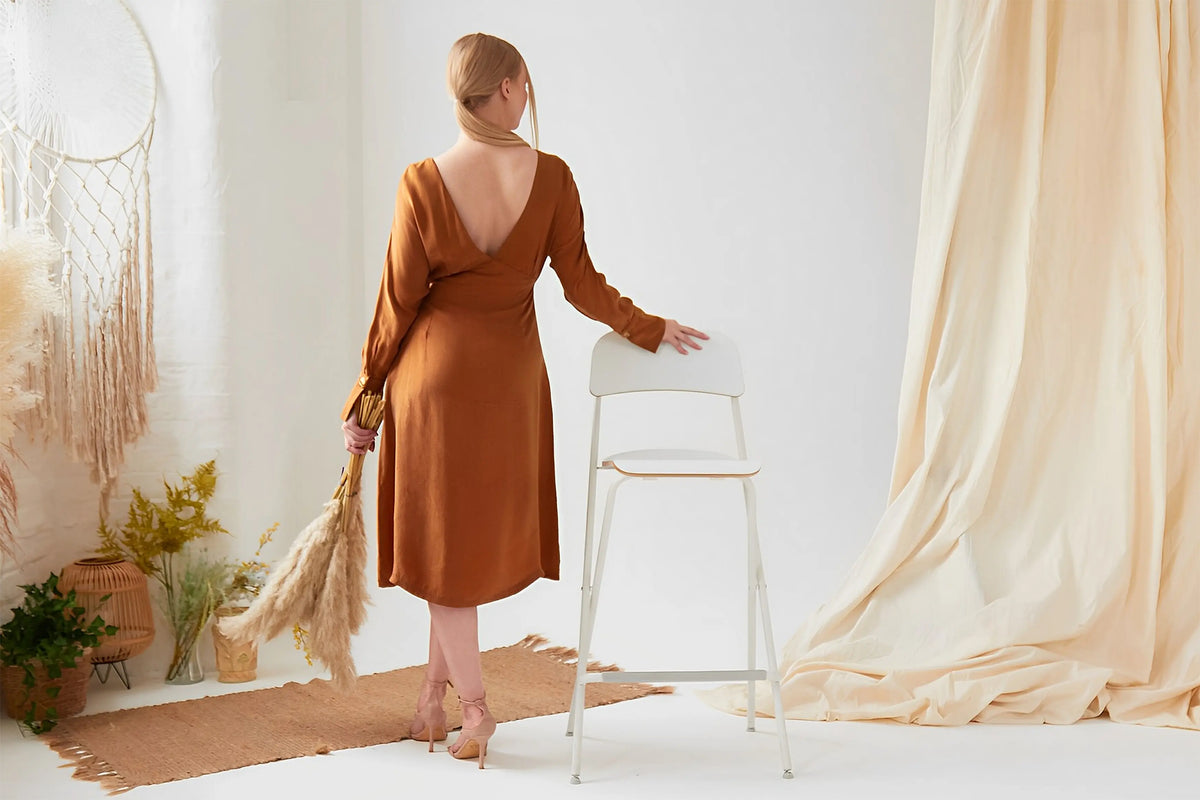 A woman in a Sarvin Mustard Yellow Midi Dress stands with her back to the camera, holding pampas grass and resting her hand on a white chair in a softly decorated, neutral-toned room.