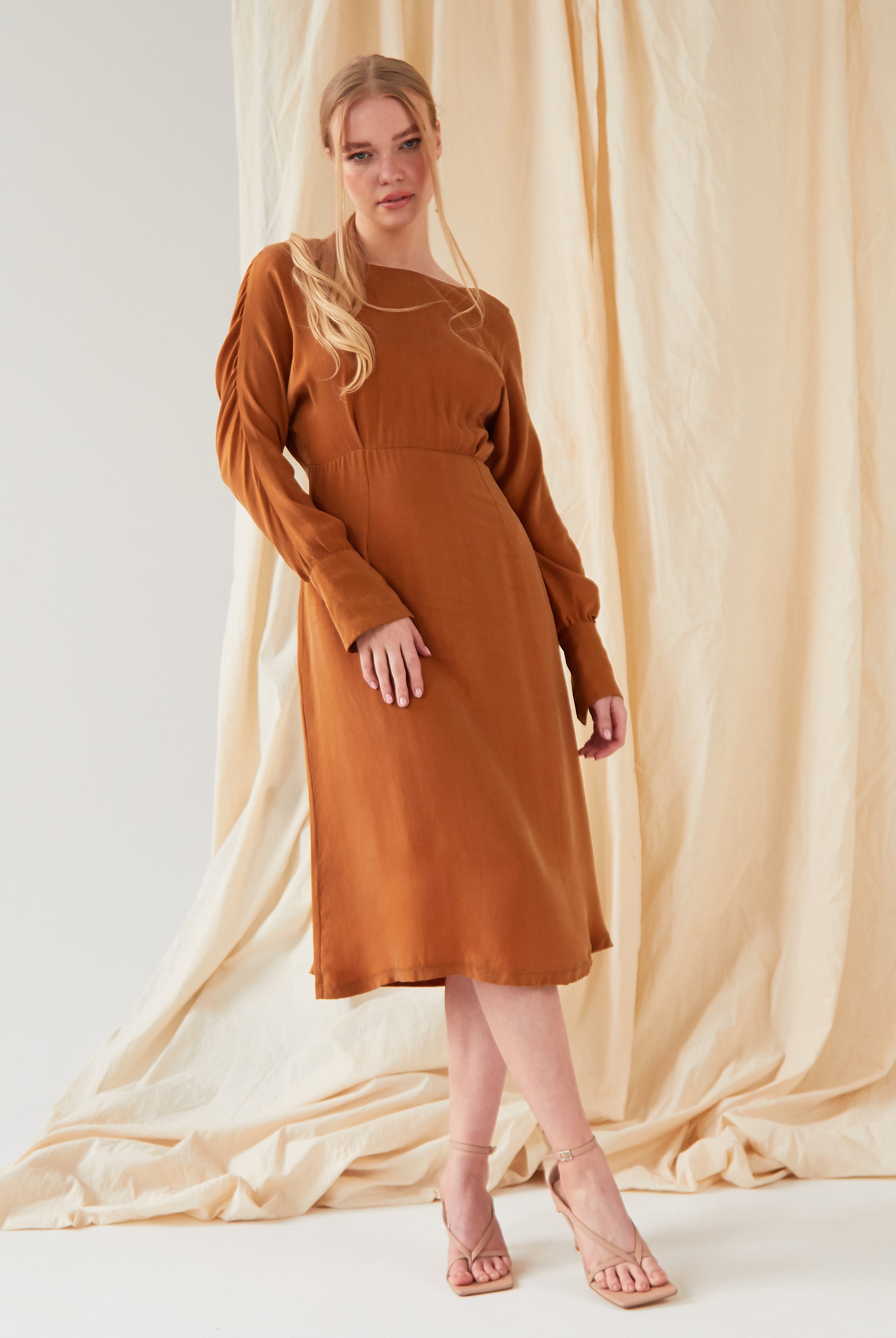 A woman with long blonde hair in a loose ponytail wears the Sarvin Mustard Yellow Midi Dress, styled with beige heeled sandals as she stands gracefully before a cream-colored draped fabric backdrop.