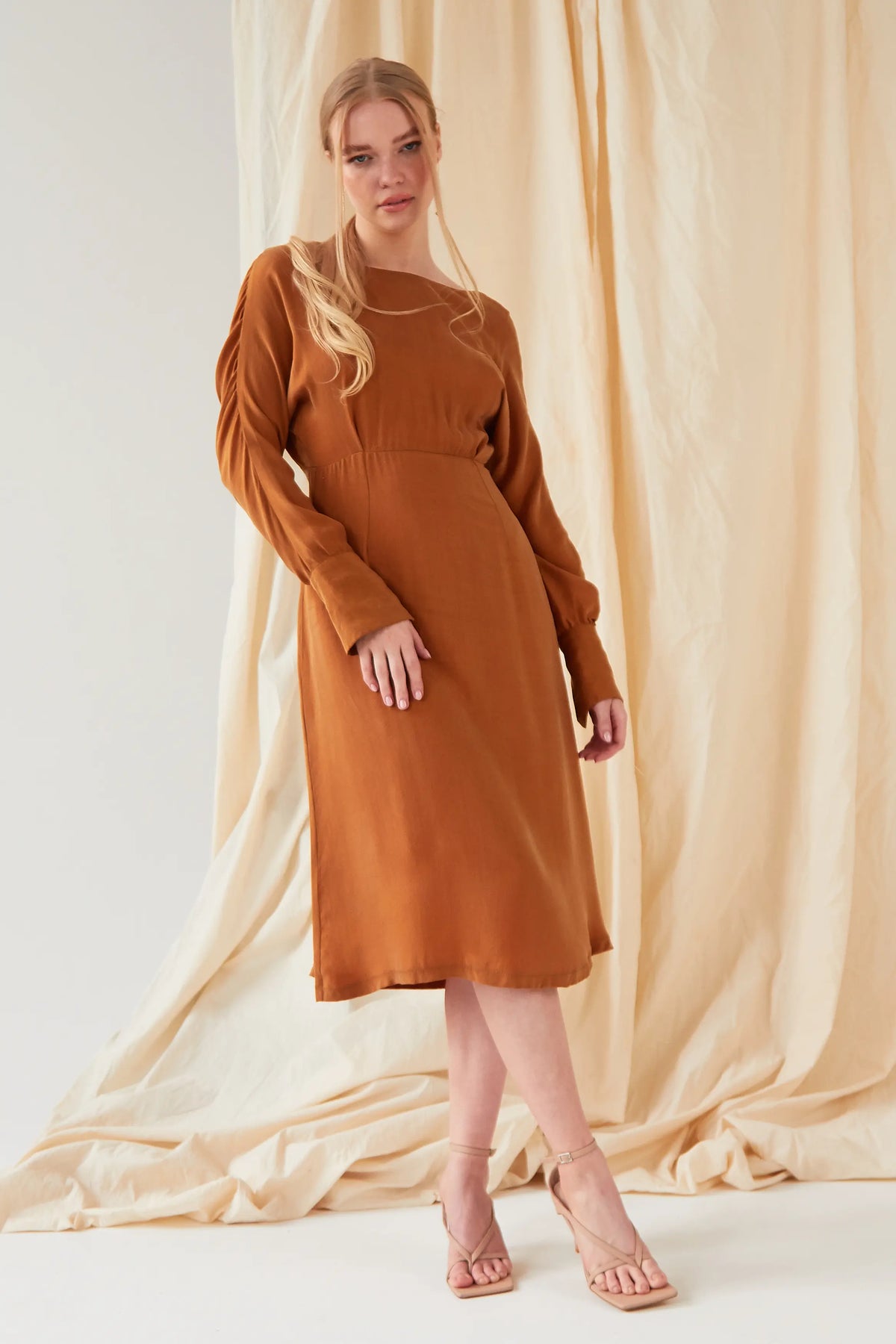 A woman with long blonde hair in a loose ponytail wears the Sarvin Mustard Yellow Midi Dress, styled with beige heeled sandals as she stands gracefully before a cream-colored draped fabric backdrop.