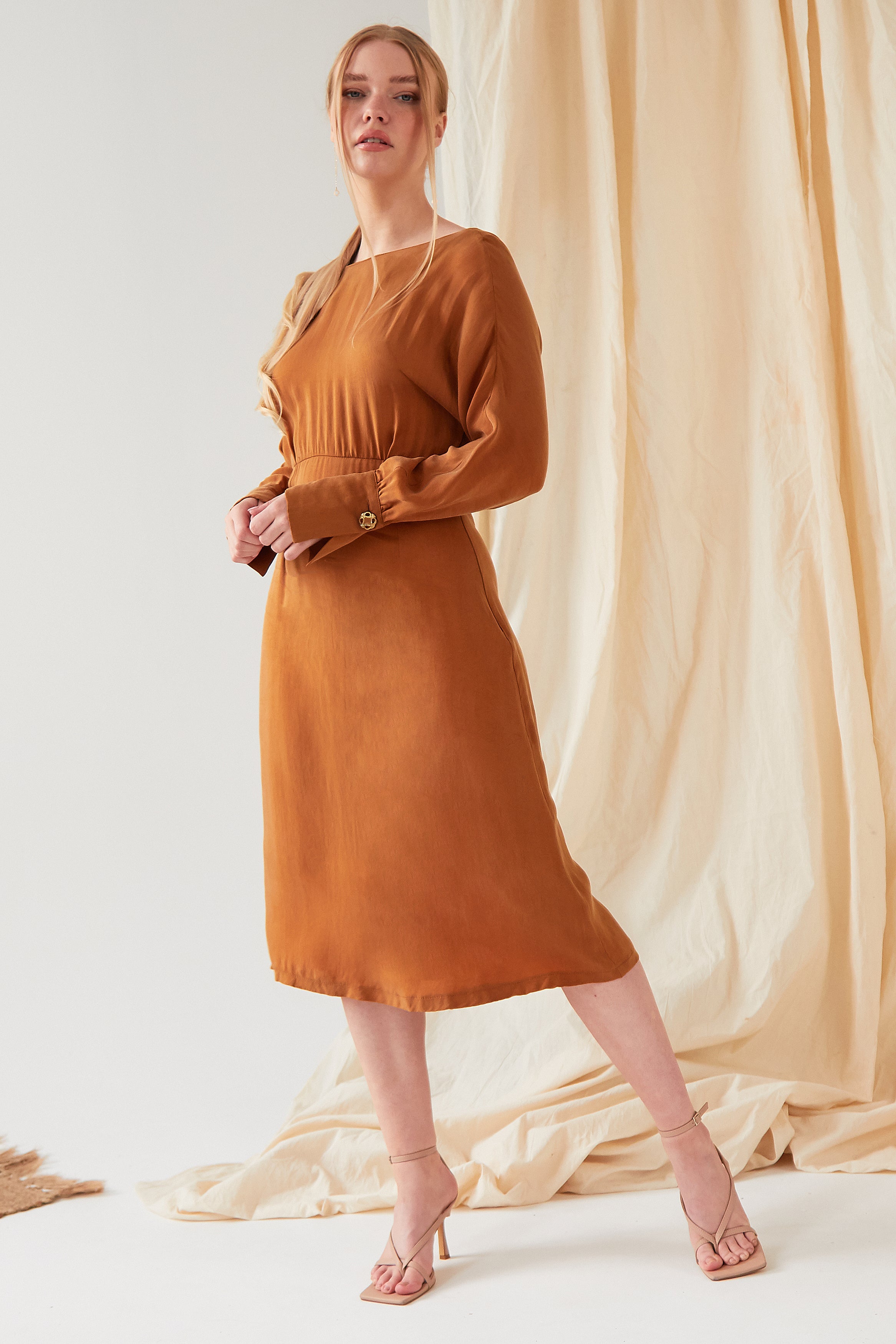 A woman wearing the Sarvin Mustard Yellow Midi Dress stands with one leg slightly bent in front of a light-colored draped fabric backdrop.