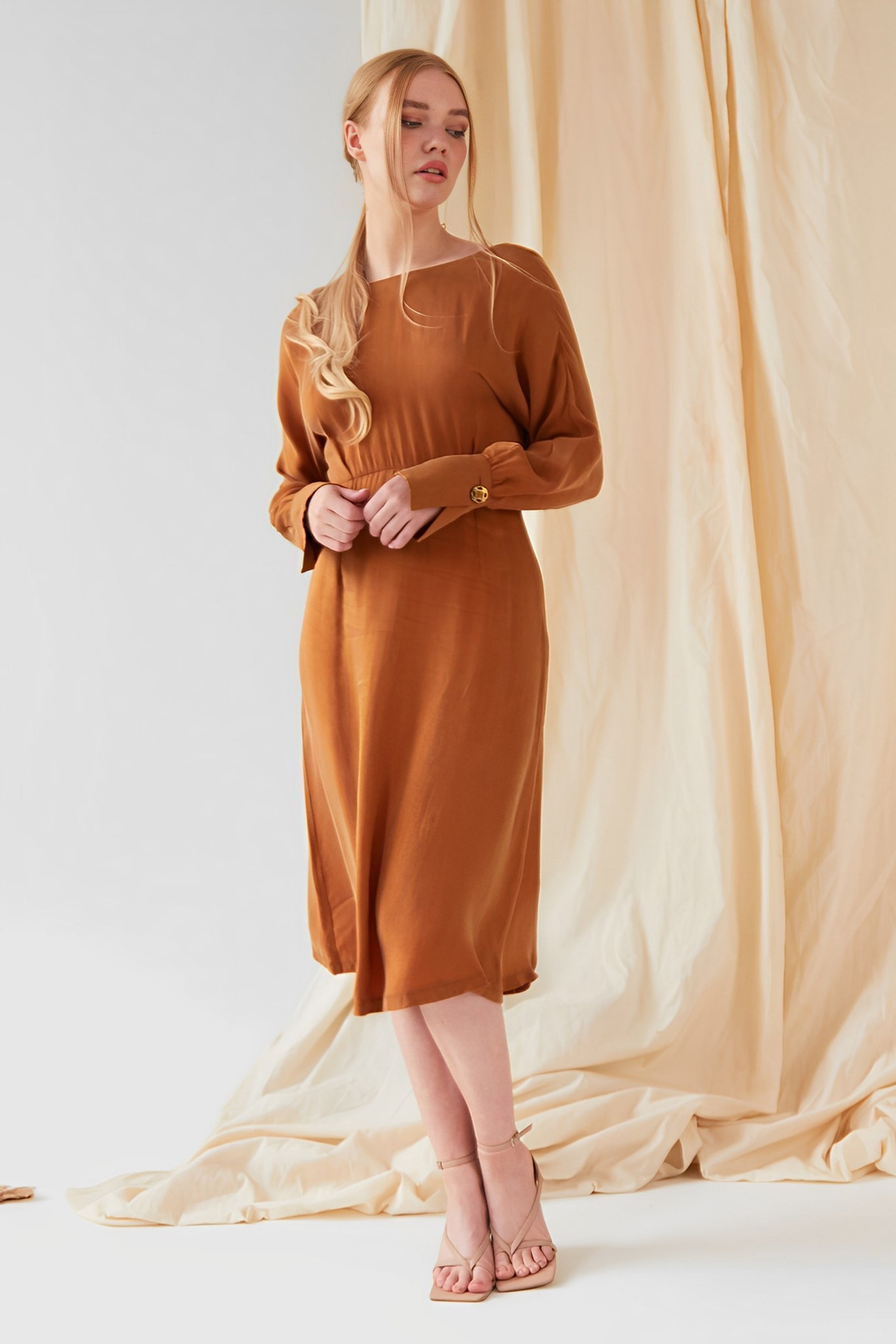 A woman stands before beige draped fabric in the Sarvin Mustard Yellow Midi Dress, paired with nude heeled sandals. Her long blonde hair is styled in a loose ponytail as she gazes slightly to the side.