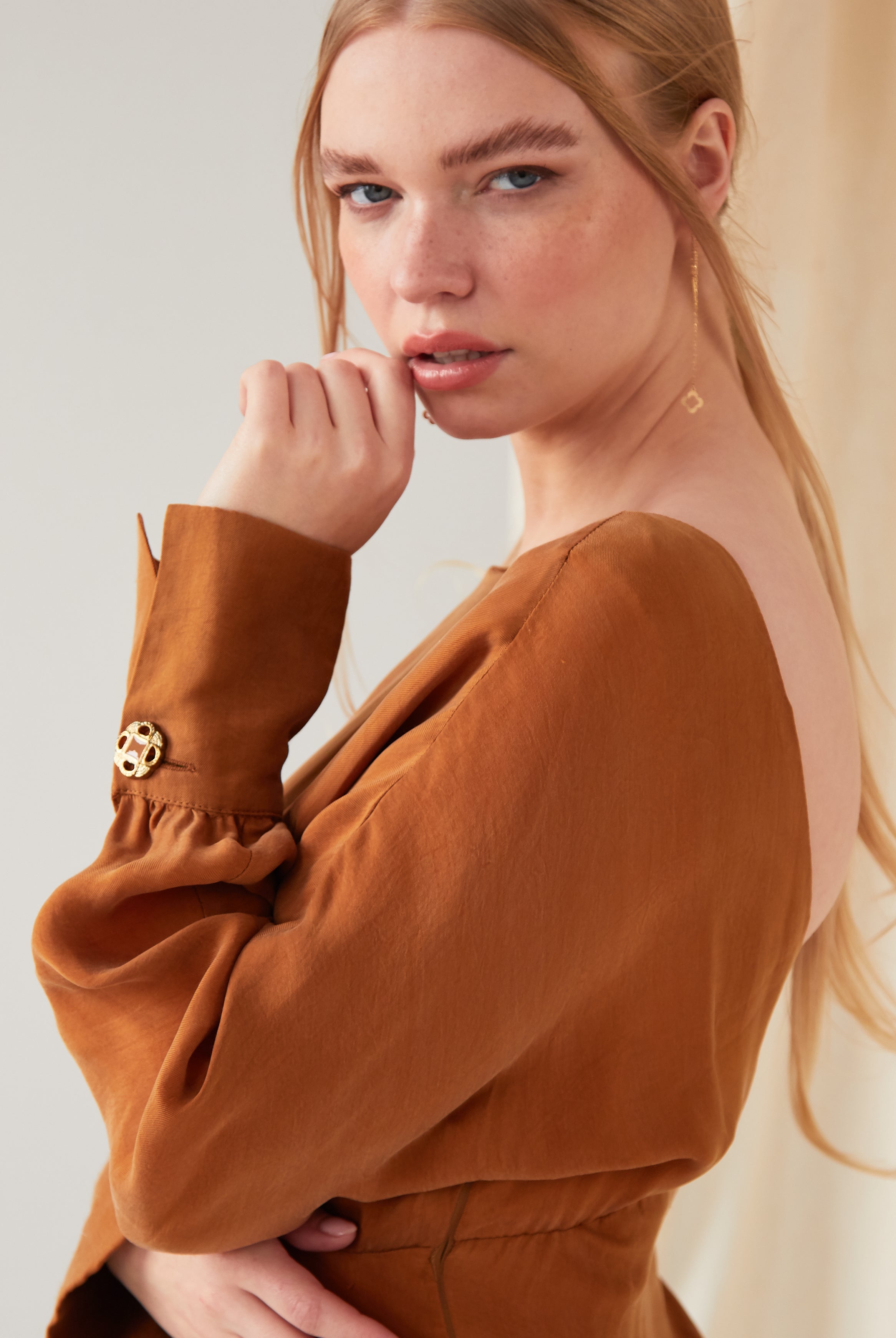 A woman with long blonde hair wears Sarvin’s rust-brown open-back top with gold buttoned sleeves and Clover Shape 18K Gold Threader Earrings. She gazes at the camera with a neutral expression, her hand near her face.