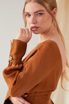 A woman with long blonde hair wears Sarvin’s rust-brown open-back top with gold buttoned sleeves and Clover Shape 18K Gold Threader Earrings. She gazes at the camera with a neutral expression, her hand near her face.