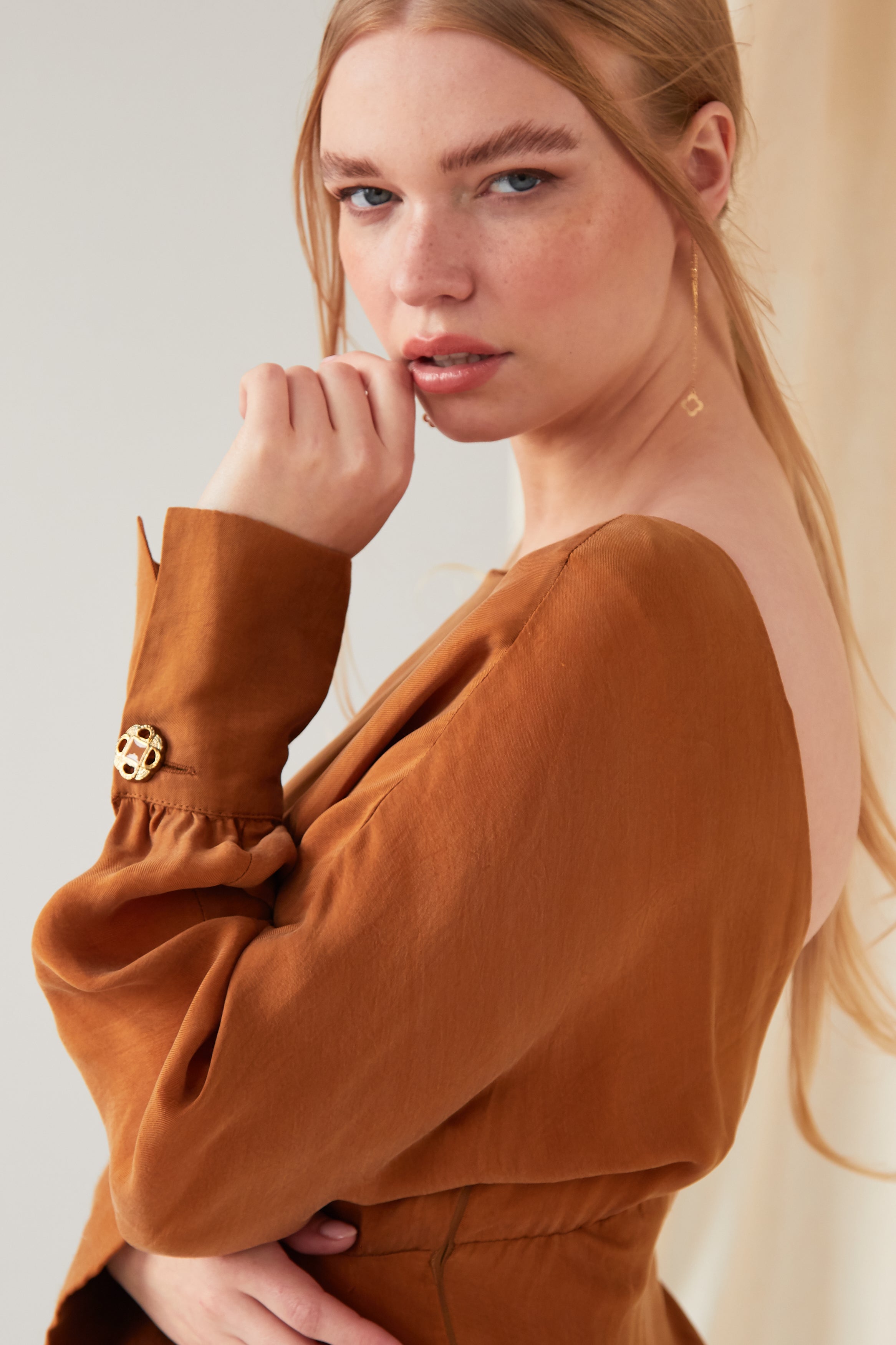 A woman with long blonde hair wears Sarvin’s rust-brown open-back top with gold buttoned sleeves and Clover Shape 18K Gold Threader Earrings. She gazes at the camera with a neutral expression, her hand near her face.
