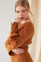 A person with long blond hair, dressed in the Sarvin Mustard Yellow Midi Dress, stands before a light background, resting one hand on their chin and the other across their waist.