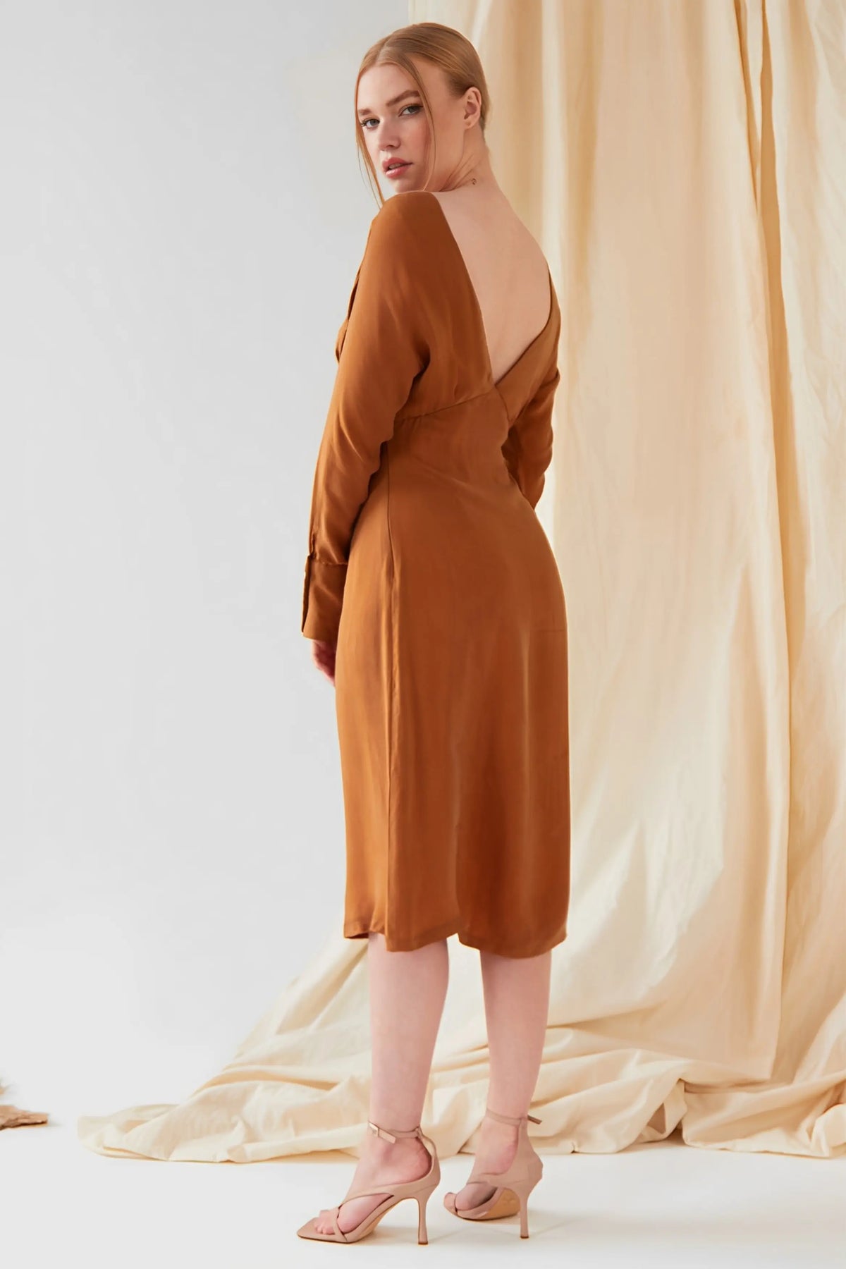 A woman wears the Sarvin Mustard Yellow Midi Dress with long sleeves and an open back, paired with light beige heels. She faces away, glancing over her shoulder against beige draped fabric.