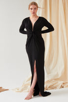Indoors, a person wears the Sarvin Sparkly Black Twist Front Dress—a maxi with long sleeves, deep V-neckline, twist knot detail, and high slit—paired with beige high-heeled sandals against a beige fabric backdrop on a white floor.