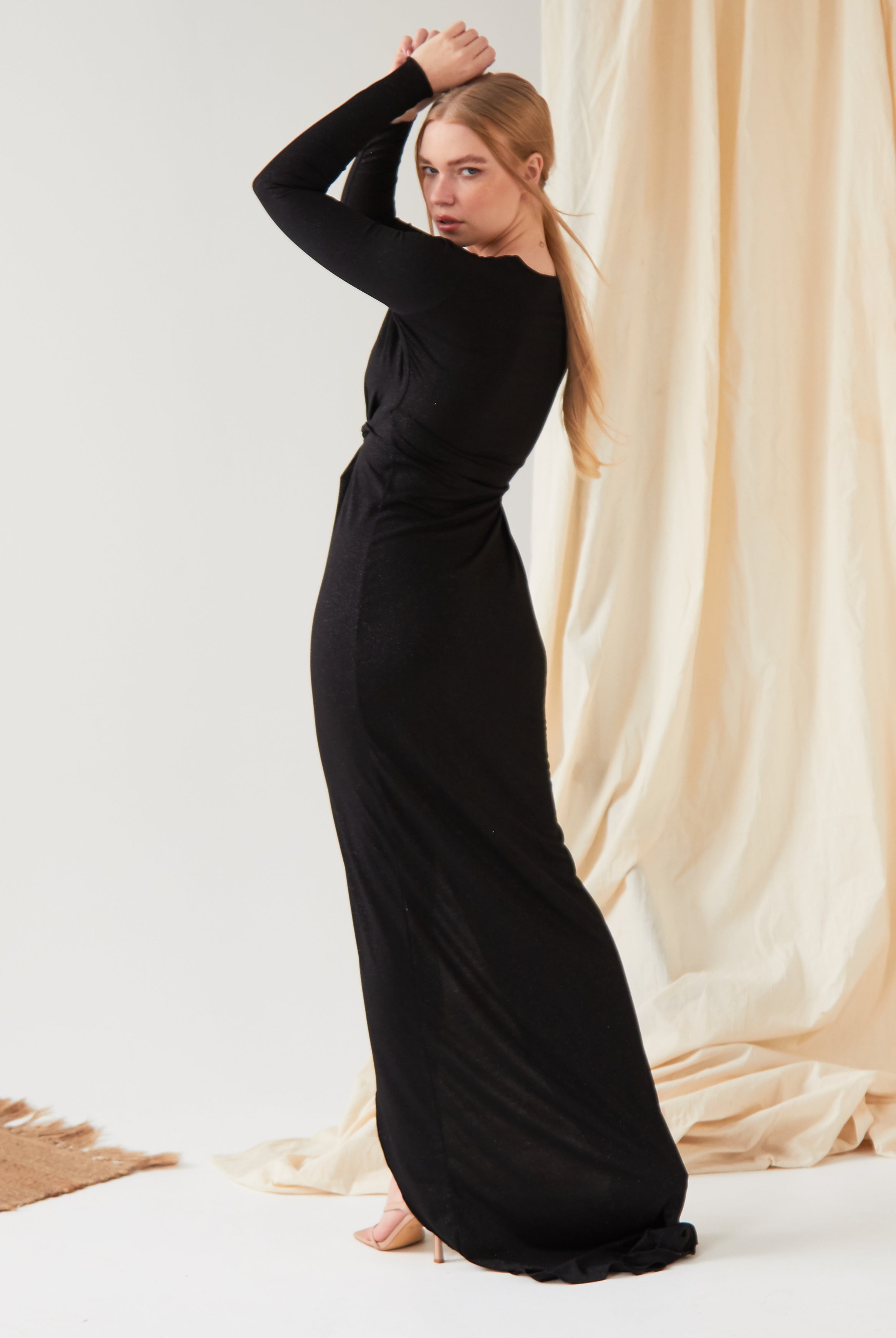 A person with long blonde hair poses sideways, arms raised above their head, in front of a beige fabric backdrop. They are wearing the Sarvin Sparkly Black Twist Front Dress on a light-colored floor.