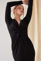 A person poses with arms raised and head tilted up, wearing the Sarvin Sparkly Black Twist Front Dress in front of a light-colored curtain.