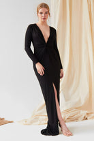 A woman stands before draped beige fabric wearing the Sarvin Sparkly Black Twist Front Dress, featuring a deep V-neck, long sleeves, and front slit, paired with nude high-heeled sandals.