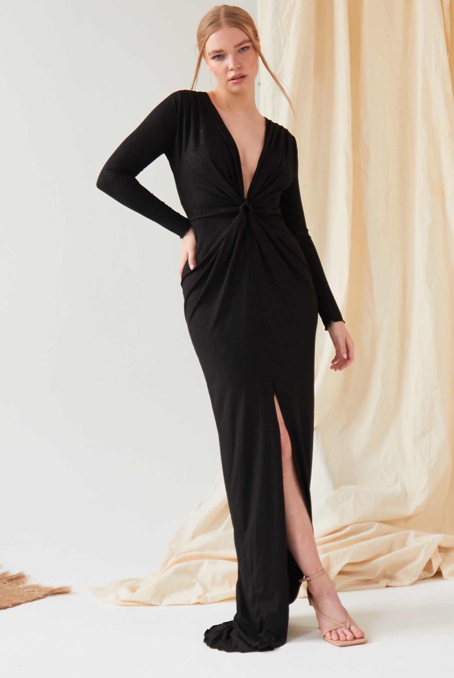 A woman poses indoors in front of beige draped fabric, wearing the Sarvin Sparkly Black Twist Front Dress—a maxi dress with a deep V-neck, long sleeves, twist knot front, and high slit.