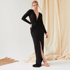 A woman poses indoors in front of beige draped fabric, wearing the Sarvin Sparkly Black Twist Front Dress—a maxi dress with a deep V-neck, long sleeves, twist knot front, and high slit.