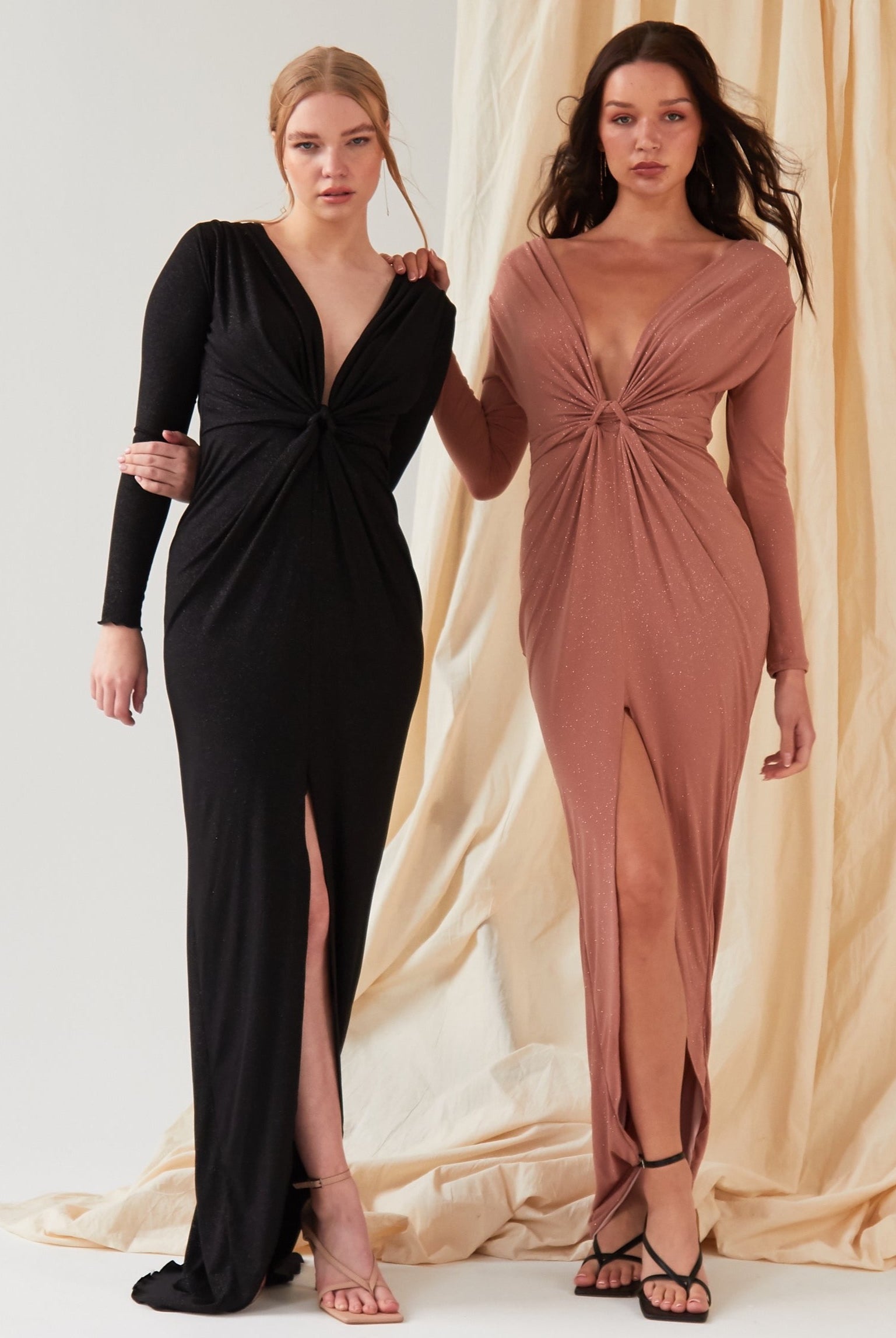 Two women wear Sarvin gowns: one in a black, long-sleeve floor-length dress with a twist knot front, deep V-neck, and thigh-high slit; the other in the Rose Gold Twist Knot Front Dress featuring the same elegant details.