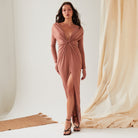 A woman stands indoors wearing Sarvin’s Rose Gold Twist Knot Front Dress with a deep V-neckline and high slit. She pairs the long-sleeved gown with black strappy heels, loose hair, and stands before neutral drapery.