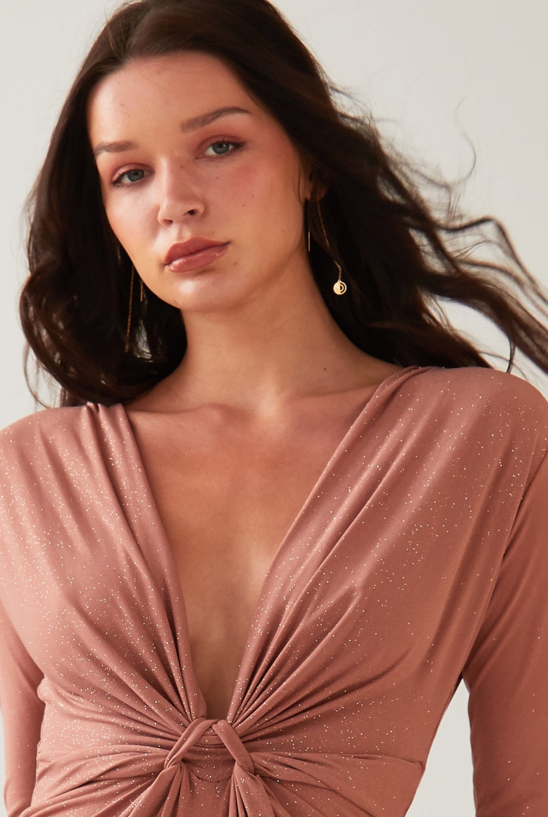 A woman with long brown hair wears a mauve, deep V-neck dress with a front knot and subtle shimmer by Sarvin. She accessorizes with Circle Shape 18K Gold Drop Earrings, standing against a plain light background.