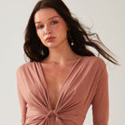 A woman with long brown hair wears a mauve, deep V-neck dress with a front knot and subtle shimmer by Sarvin. She accessorizes with Circle Shape 18K Gold Drop Earrings, standing against a plain light background.