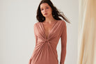 A woman wears the Sarvin Rose Gold Twist Knot Front Dress with long sleeves and a deep V-neckline, standing before a plain background, a sheer curtain visible to her right.