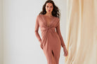 A woman wears the Sarvin Rose Gold Twist Knot Front Dress with long sleeves and a high slit, standing indoors against light-colored curtains.