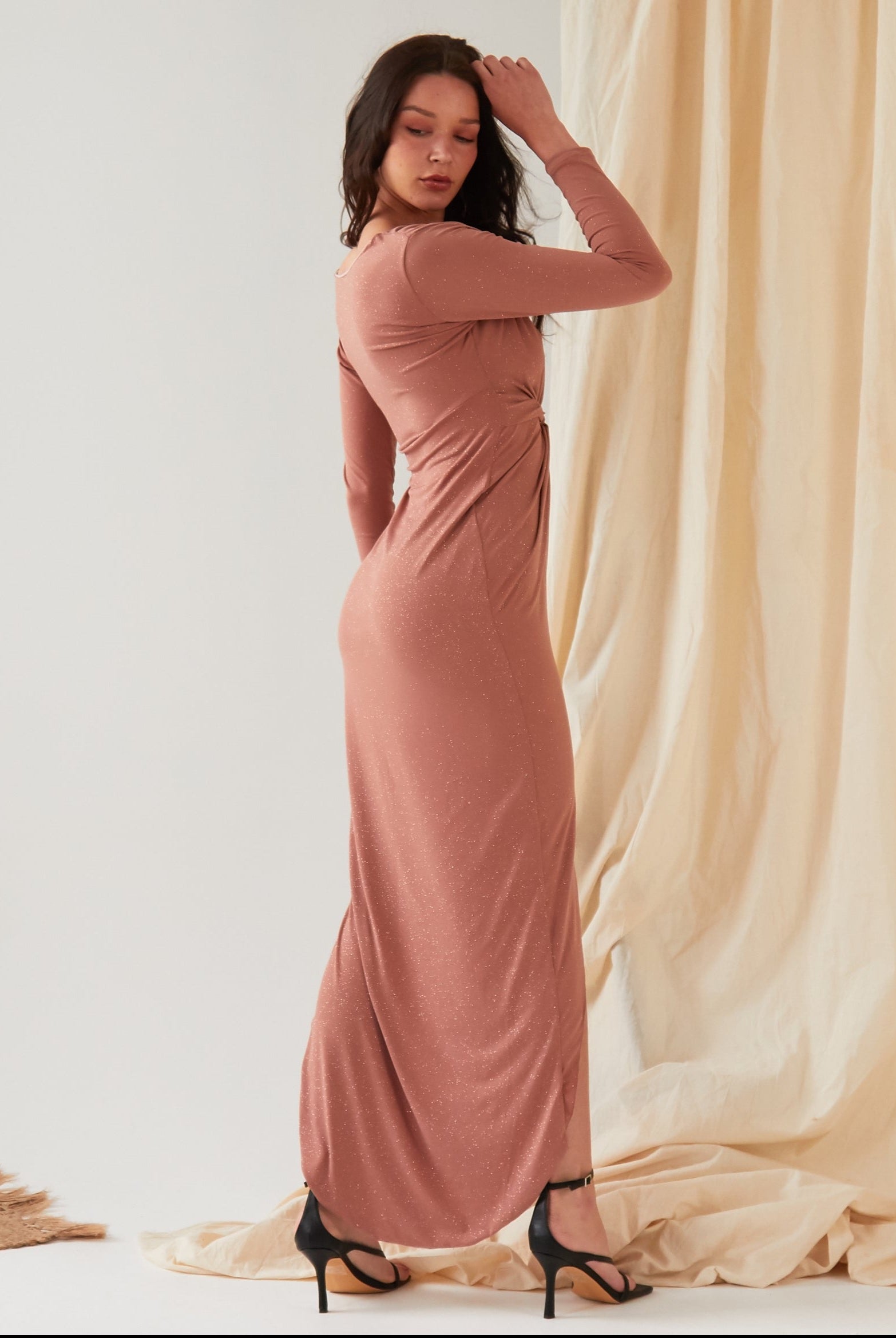 A woman stands sideways against draped fabric, wearing the Sarvin Rose Gold Twist Knot Front Dress with long sleeves and a fitted silhouette, paired with black high-heeled sandals and one hand touching her head.