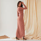 A woman stands sideways against draped fabric, wearing the Sarvin Rose Gold Twist Knot Front Dress with long sleeves and a fitted silhouette, paired with black high-heeled sandals and one hand touching her head.