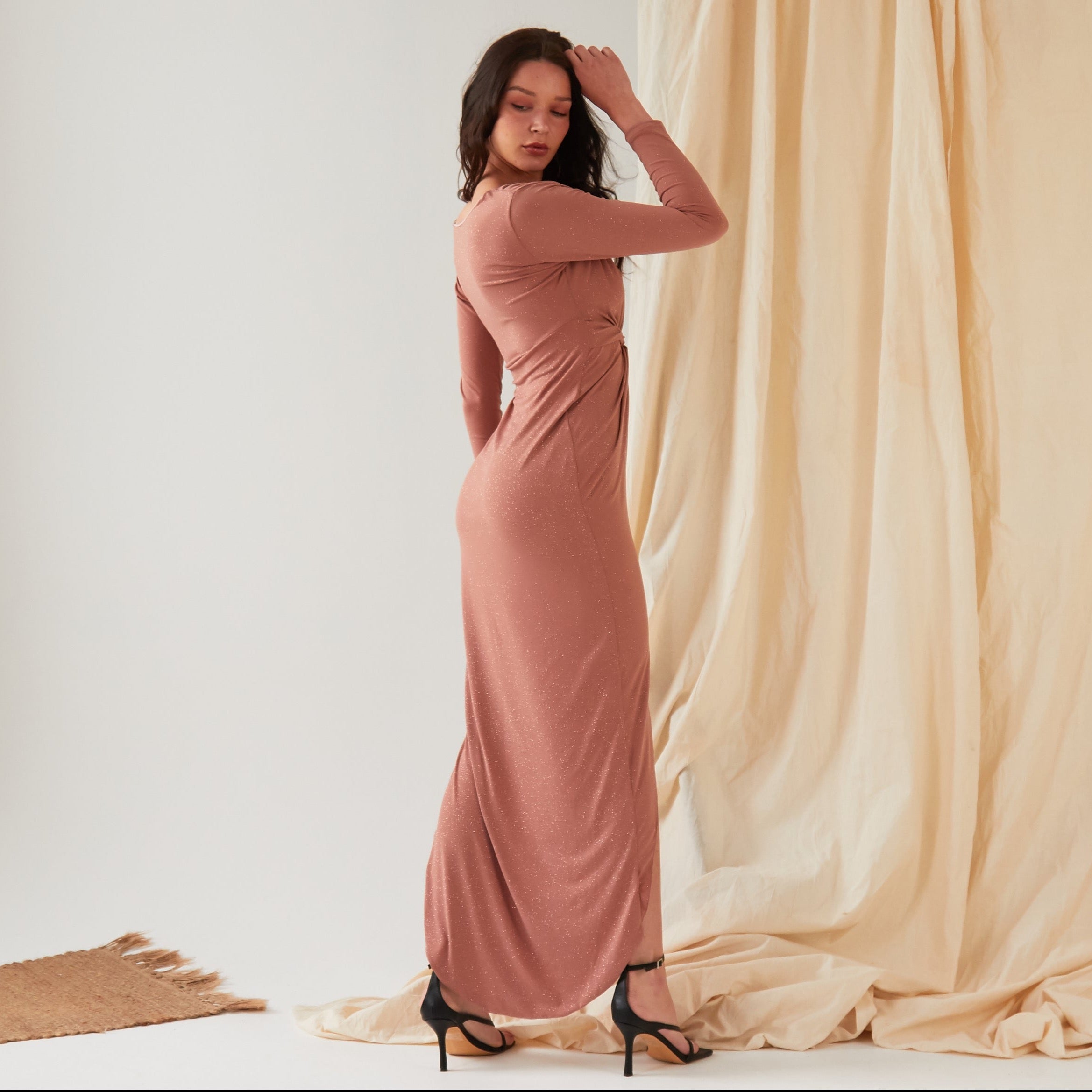 A woman stands sideways against draped fabric, wearing the Sarvin Rose Gold Twist Knot Front Dress with long sleeves and a fitted silhouette, paired with black high-heeled sandals and one hand touching her head.