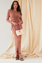 A woman stands before beige drapes wearing the Sarvin Rose Gold Twist Knot Front Dress featuring long sleeves and a deep V-neck, styled with a cream handbag adorned with pink beads and black strappy sandals.