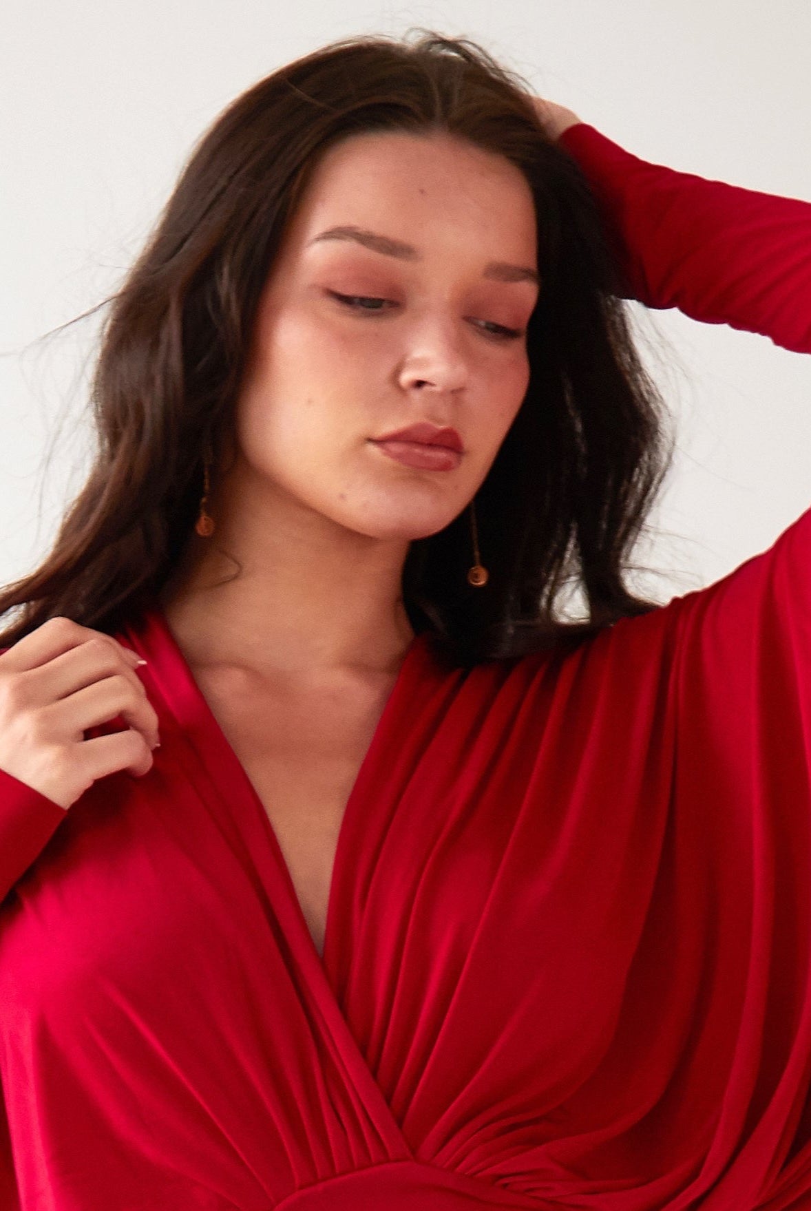 A woman with long dark hair wears Sarvin’s Circle Shape 18K Gold Drop Earrings and a red, long-sleeved draped dress. Posing against a plain light background, she gazes down with one hand near her head and the other at her shoulder.
