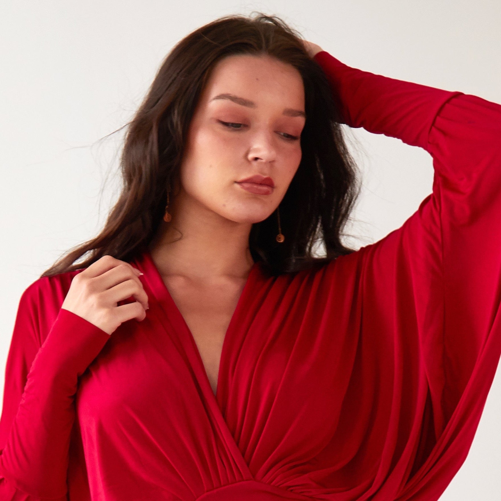 A woman with long dark hair wears Sarvin’s Circle Shape 18K Gold Drop Earrings and a red, long-sleeved draped dress. Posing against a plain light background, she gazes down with one hand near her head and the other at her shoulder.