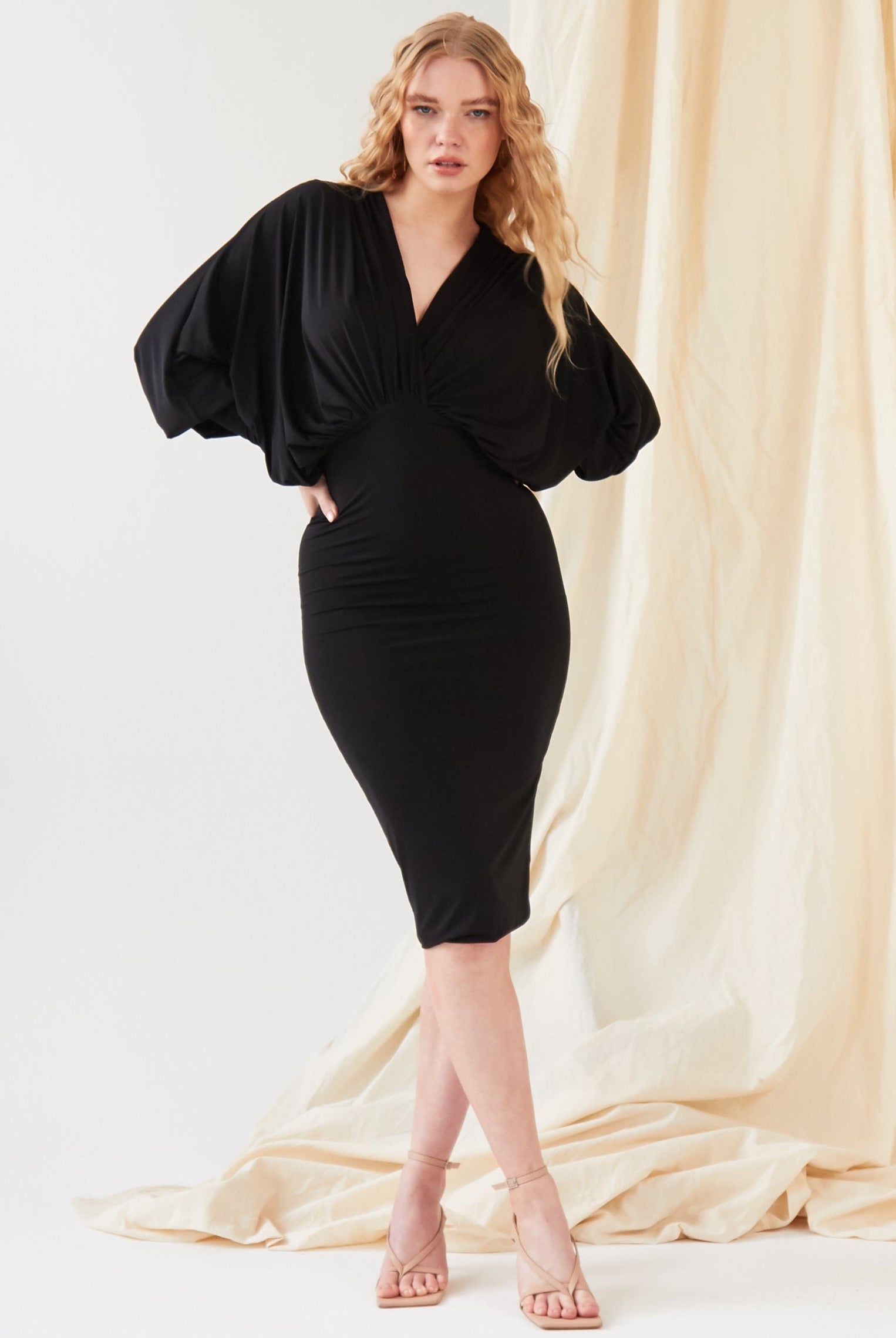 A woman poses before a beige draped backdrop in the Sarvin Black Batwing Sleeve Midi Dress, featuring silk-touch jersey. She pairs the look with nude-heeled sandals and lets her long wavy blonde hair frame her neutral expression.