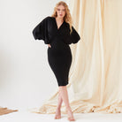 A woman poses before a beige draped backdrop in the Sarvin Black Batwing Sleeve Midi Dress, featuring silk-touch jersey. She pairs the look with nude-heeled sandals and lets her long wavy blonde hair frame her neutral expression.