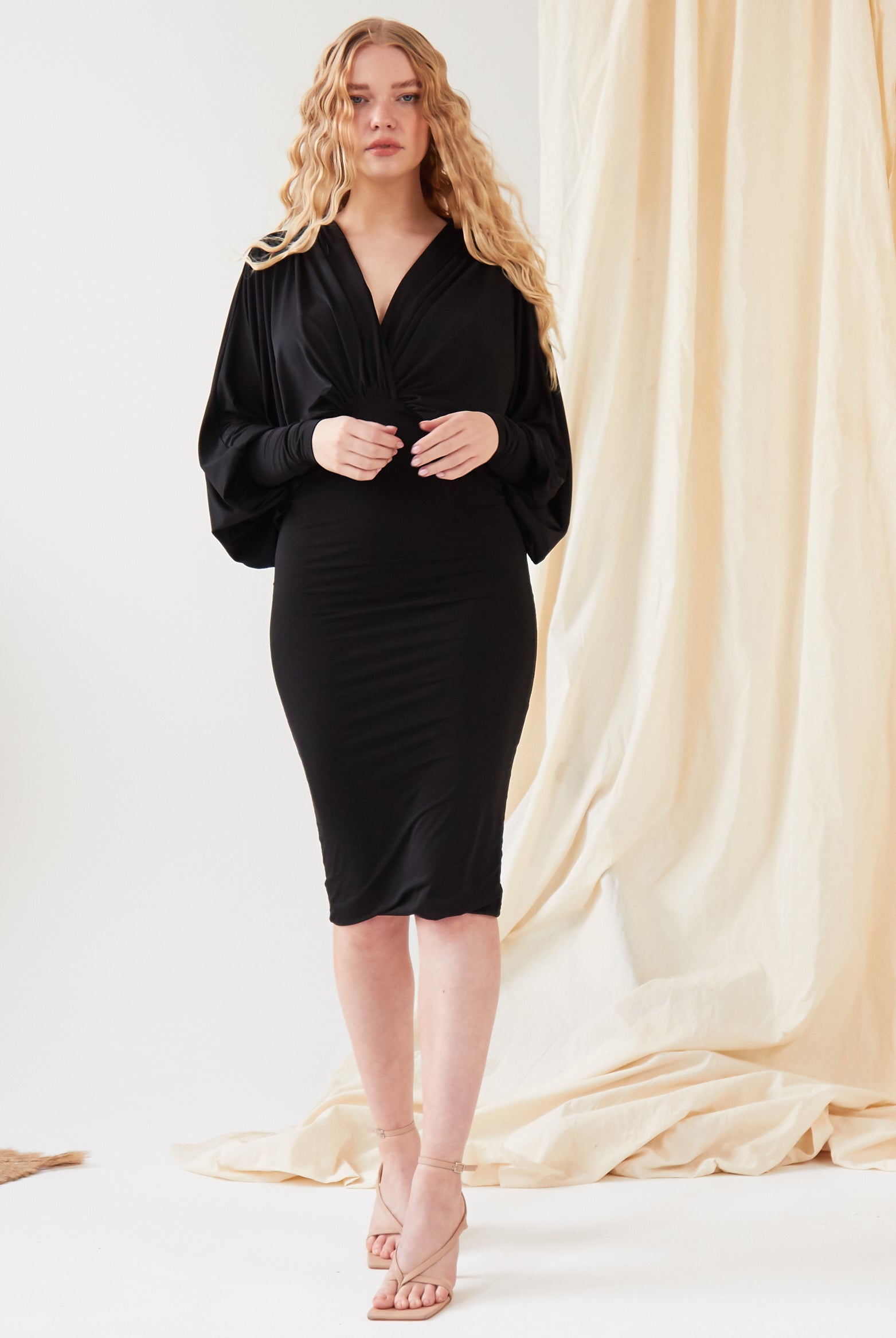 A person with long, wavy blond hair stands indoors wearing the Sarvin Black Batwing Sleeve Midi Dress and beige heels, with neutral pampas grass and light fabric drapes in the background.