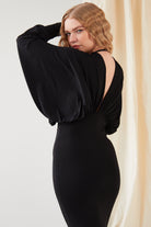 A person with long, wavy blonde hair models the Sarvin Black Batwing Sleeve Midi Dress, featuring an open back. They pose with one arm raised, gazing over their shoulder before a cream-colored curtain backdrop.