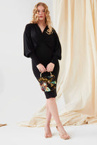 A woman with long wavy hair stands on a cream-colored floor, wearing the Sarvin Black Batwing Sleeve Midi Dress and nude heels, holding a small floral handbag. Cream fabric drapes in the background.