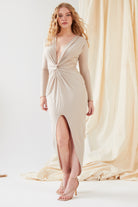 A woman indoors wears the Sarvin Low Cut Midi Dress in beige, featuring a knot front, long sleeves, deep V-neckline, and front slit. She pairs it with strappy heels; her long wavy hair flows as neutral fabric drapes softly behind her.