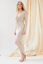 A woman models the Sarvin Low Cut Midi Dress, featuring a light beige knot front, long sleeves, and a deep V-neckline. She pairs it with nude high-heeled sandals against a cream draped fabric backdrop.