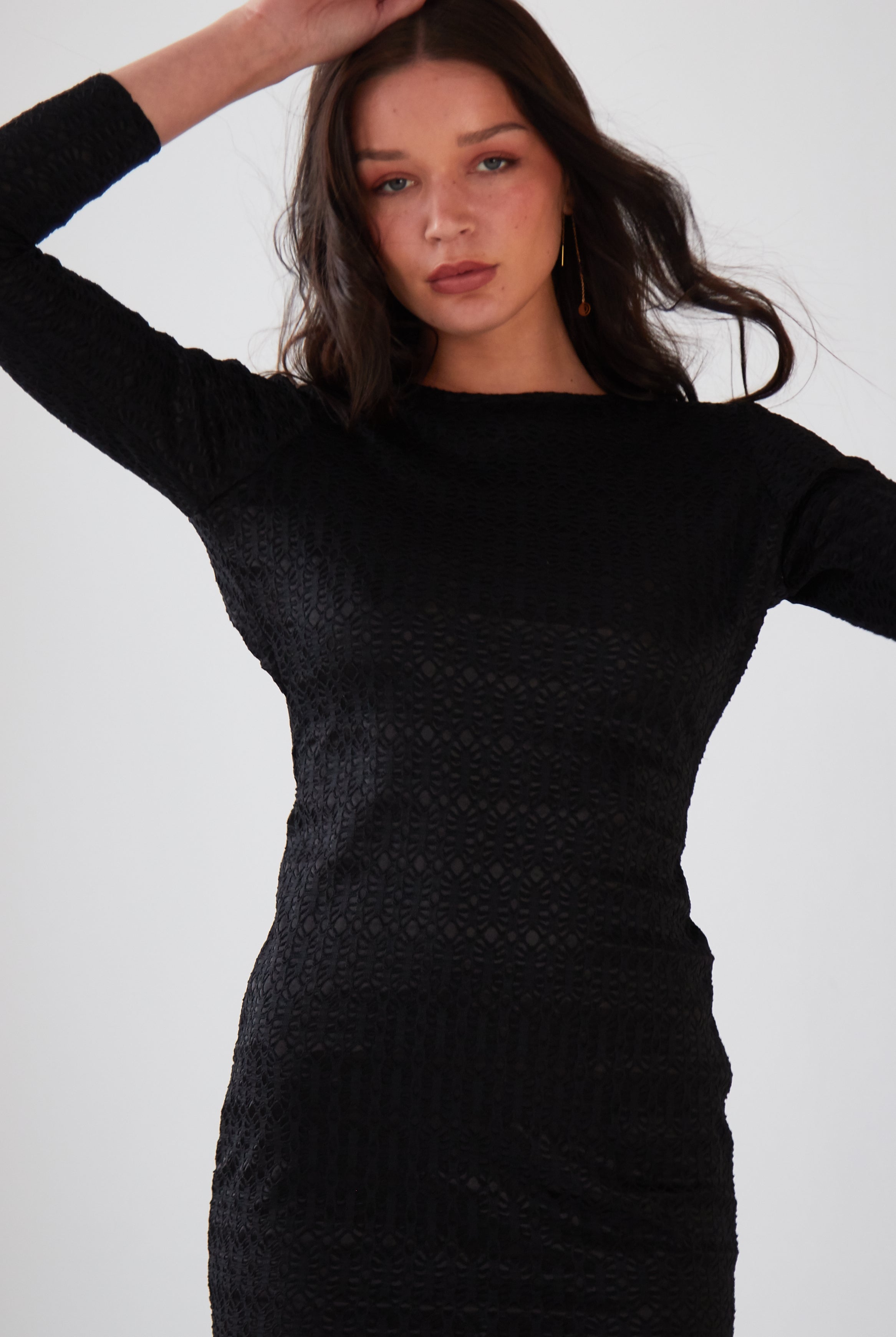 A woman with long dark hair wears the Sarvin Black Twisted Backless Dress with dangle earrings, raising one arm above her head against a plain light background.