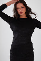 A woman with long dark hair wears the Sarvin Black Twisted Backless Dress with dangle earrings, raising one arm above her head against a plain light background.