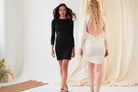 Two women stand indoors; one faces forward in a black lace bodycon dress, while the other, facing away, wears the Sarvin Black Twisted Backless Dress. Neutral fabrics and plants complete the backdrop.