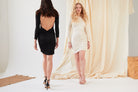 Two women pose indoors against a light fabric backdrop. One wears a white long-sleeve dress and nude heels. The other models the Sarvin Black Twisted Backless Dress, featuring a knotted detail, paired with black heels and facing away from the camera.