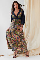 A woman indoors models the Sarvin Hand painted Long Sleeve Maxi Dress, featuring a dark blue bodice, deep V-neck, and colorful paisley-patterned skirt. She holds one side of the eco-friendly dress and pairs it with black strappy sandals.