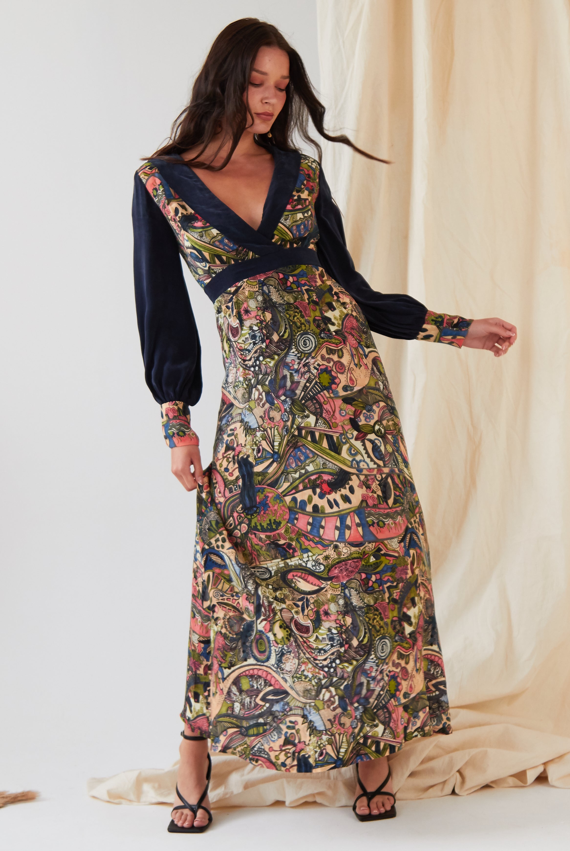 A woman poses indoors with one hand on her hip, wearing the Sarvin Hand painted Long Sleeve Maxi Dress featuring colorful, intricate patterns and a deep V-neck, set against a light fabric backdrop.