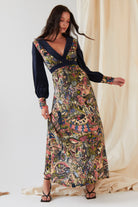 A woman poses indoors with one hand on her hip, wearing the Sarvin Hand painted Long Sleeve Maxi Dress featuring colorful, intricate patterns and a deep V-neck, set against a light fabric backdrop.