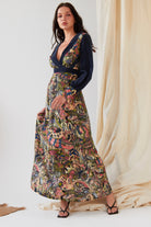 A woman with long dark hair and black strappy heels stands indoors in the Sarvin Hand painted Long Sleeve Maxi Dress, featuring a colorful paisley pattern, deep V-neck, and navy sleeves. Neutral fabric drapes in the background.