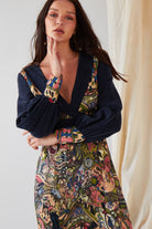 A person with long dark hair wears the Sarvin Hand Painted Long Sleeve Maxi Dress, showcasing a dark blue top with a colorful, intricate patterned skirt and cuffs. They rest a hand on their chin before a light background and beige curtain.