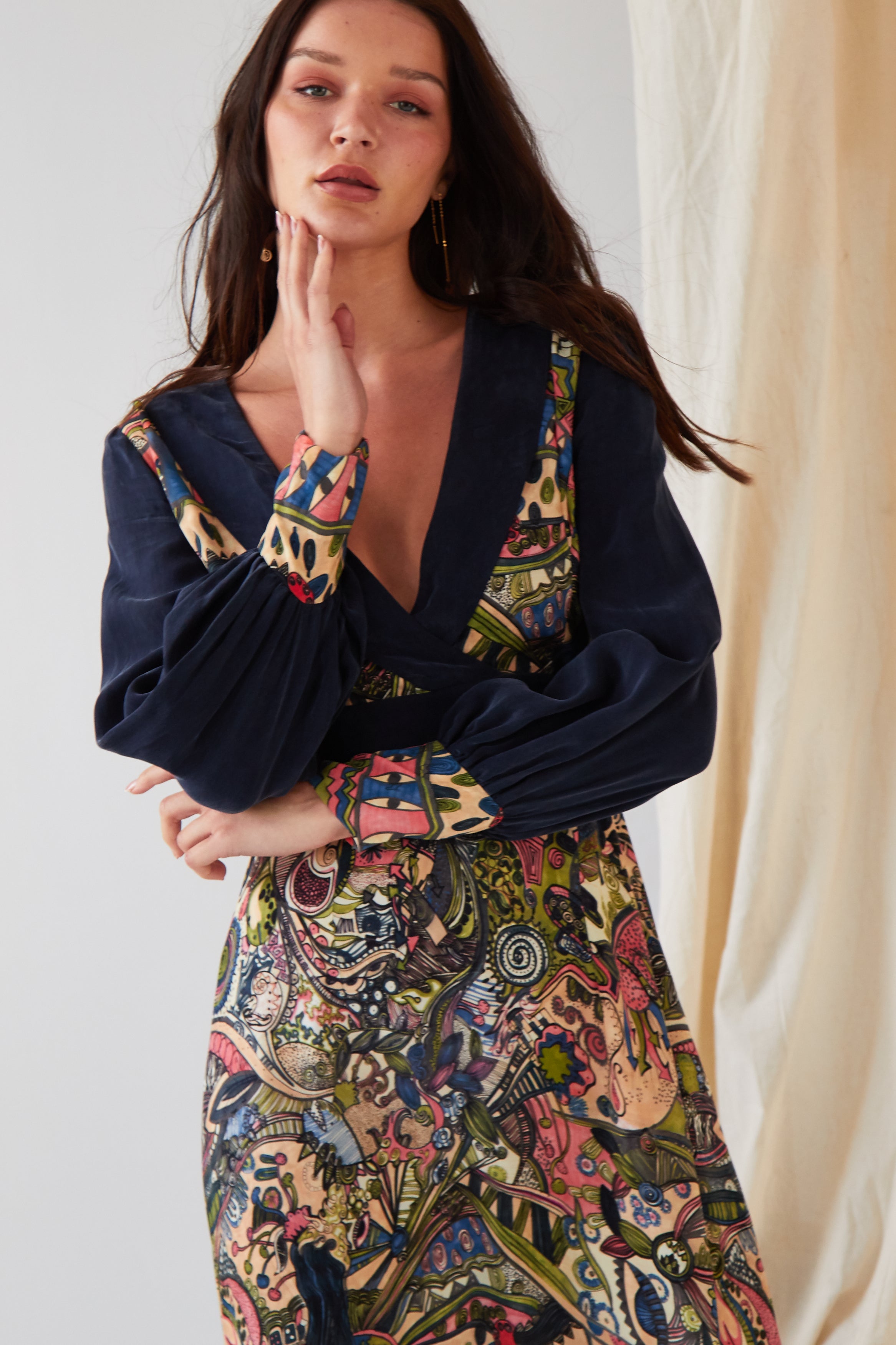 A person with long dark hair wears the Sarvin Hand Painted Long Sleeve Maxi Dress, showcasing a dark blue top with a colorful, intricate patterned skirt and cuffs. They rest a hand on their chin before a light background and beige curtain.