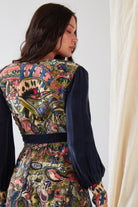 A person with long dark hair wears the Sarvin Hand painted Long Sleeve Maxi Dress, featuring intricate, colorful abstract patterns on the back and solid dark blue sleeves. They stand sideways, looking over their shoulder.
