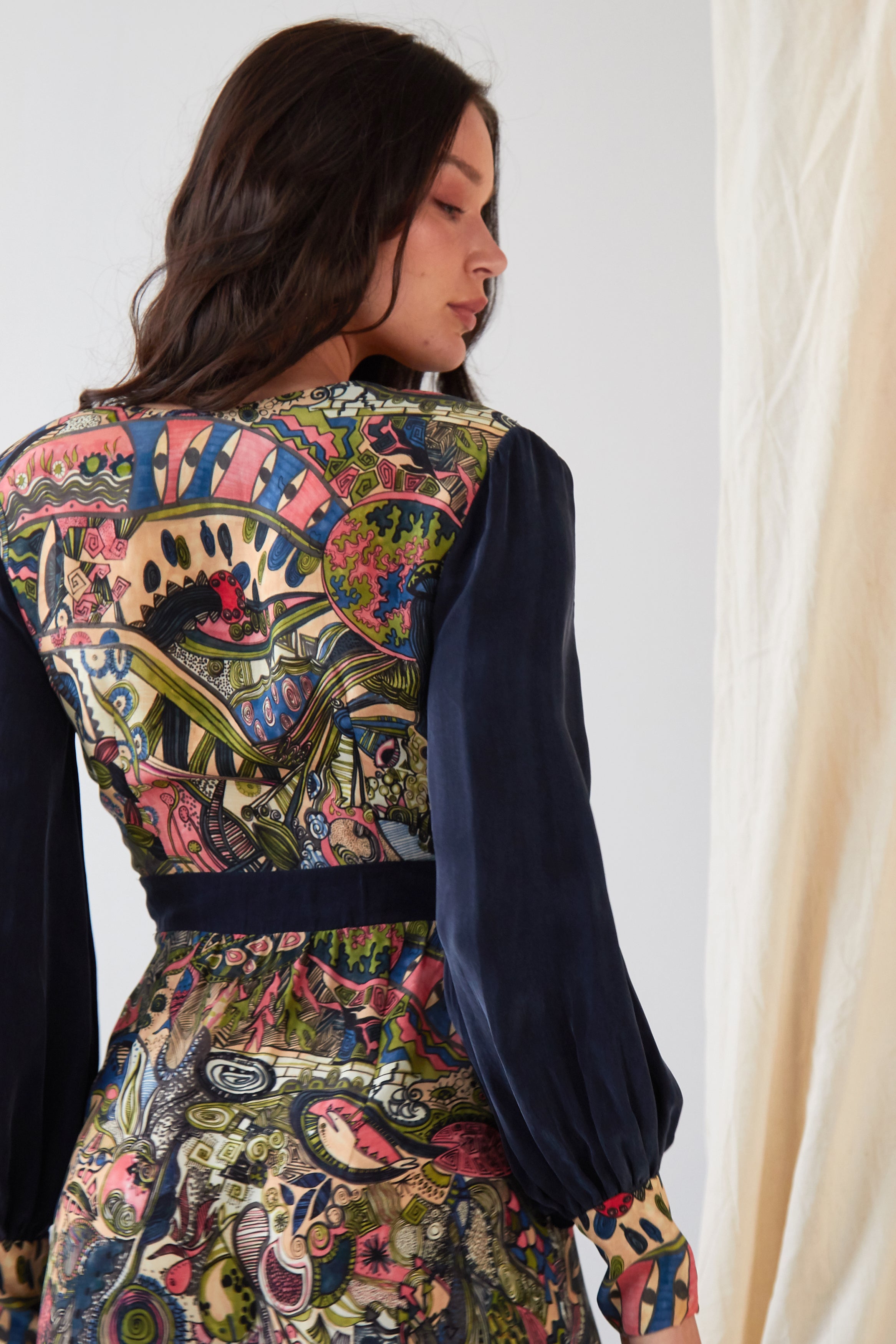 A person with long dark hair wears the Sarvin Hand painted Long Sleeve Maxi Dress, featuring intricate, colorful abstract patterns on the back and solid dark blue sleeves. They stand sideways, looking over their shoulder.