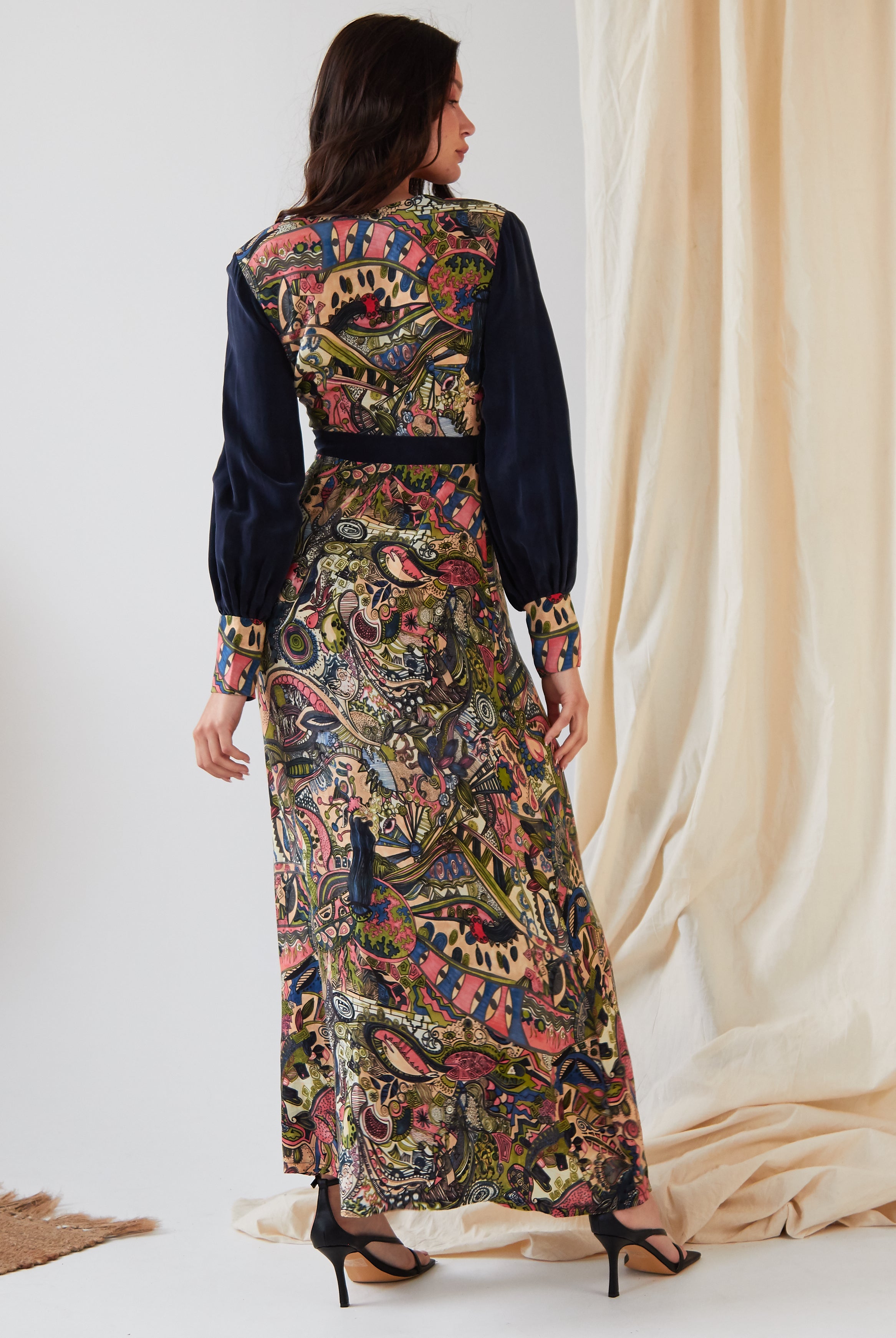 A woman faces away, wearing the Sarvin Hand Painted Long Sleeve Maxi Dress featuring a colorful paisley and floral design with dark sleeves and waistband, paired with black heels before softly draped light fabric.